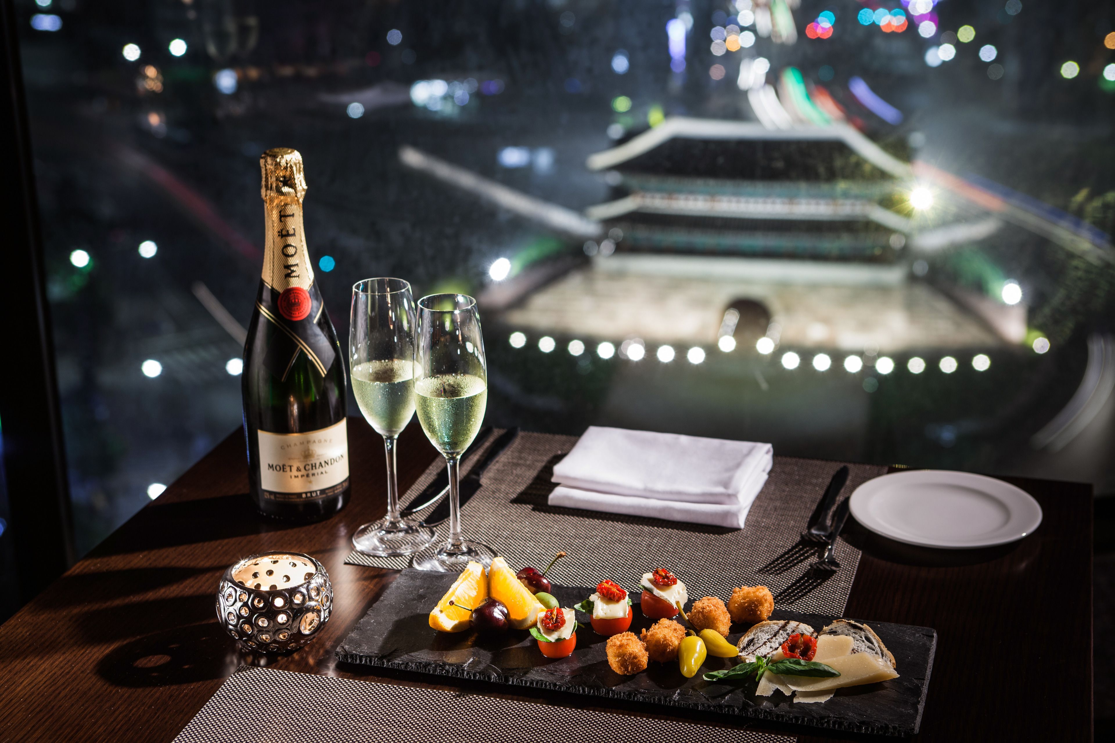 A table with a bottle of Moët & Chandon champagne, two glasses, a variety of appetizers, and a view of a traditional building through a window.