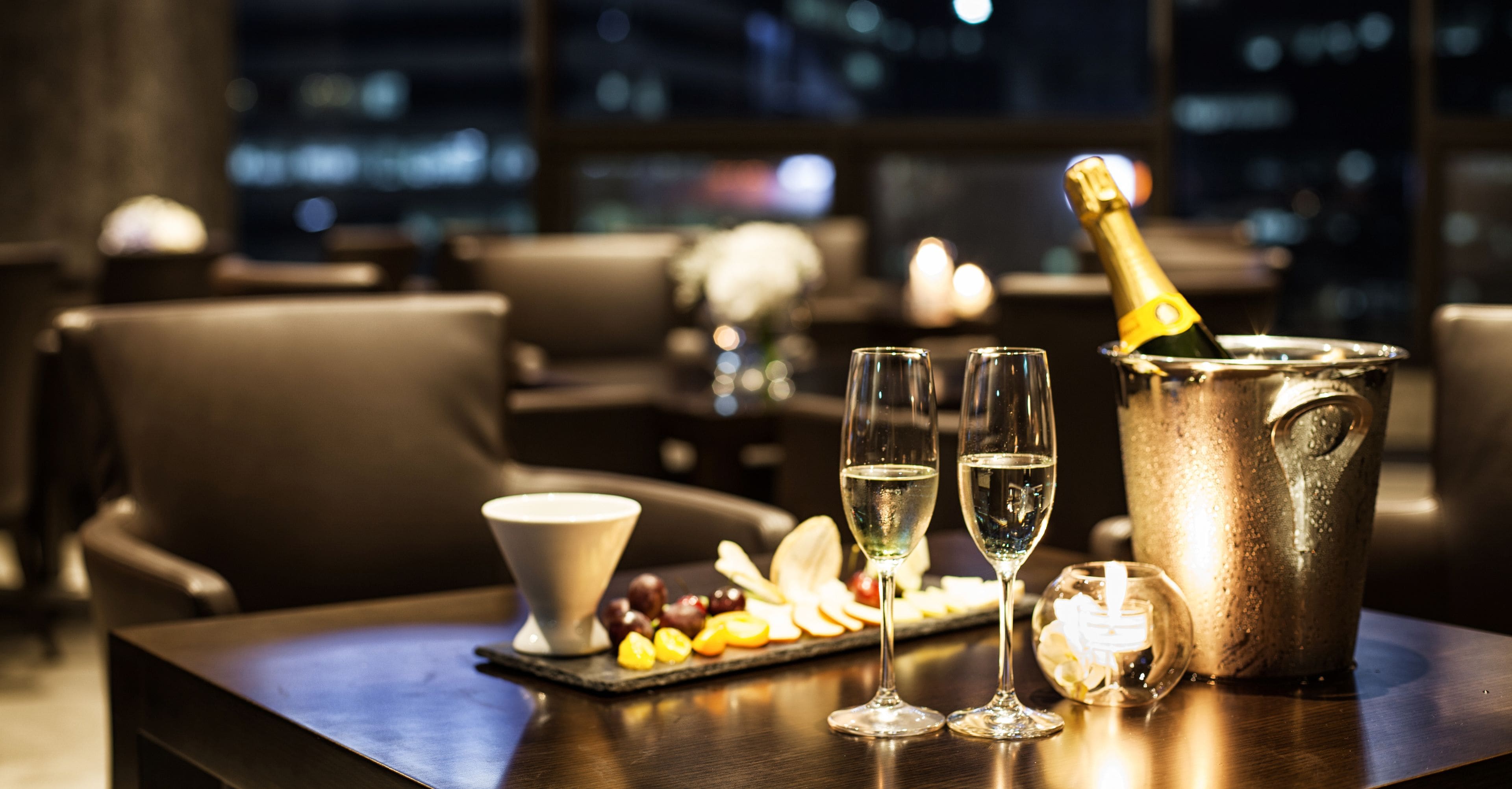 A table with a bottle of champagne, two glasses, a bowl, and a plate of fruit.