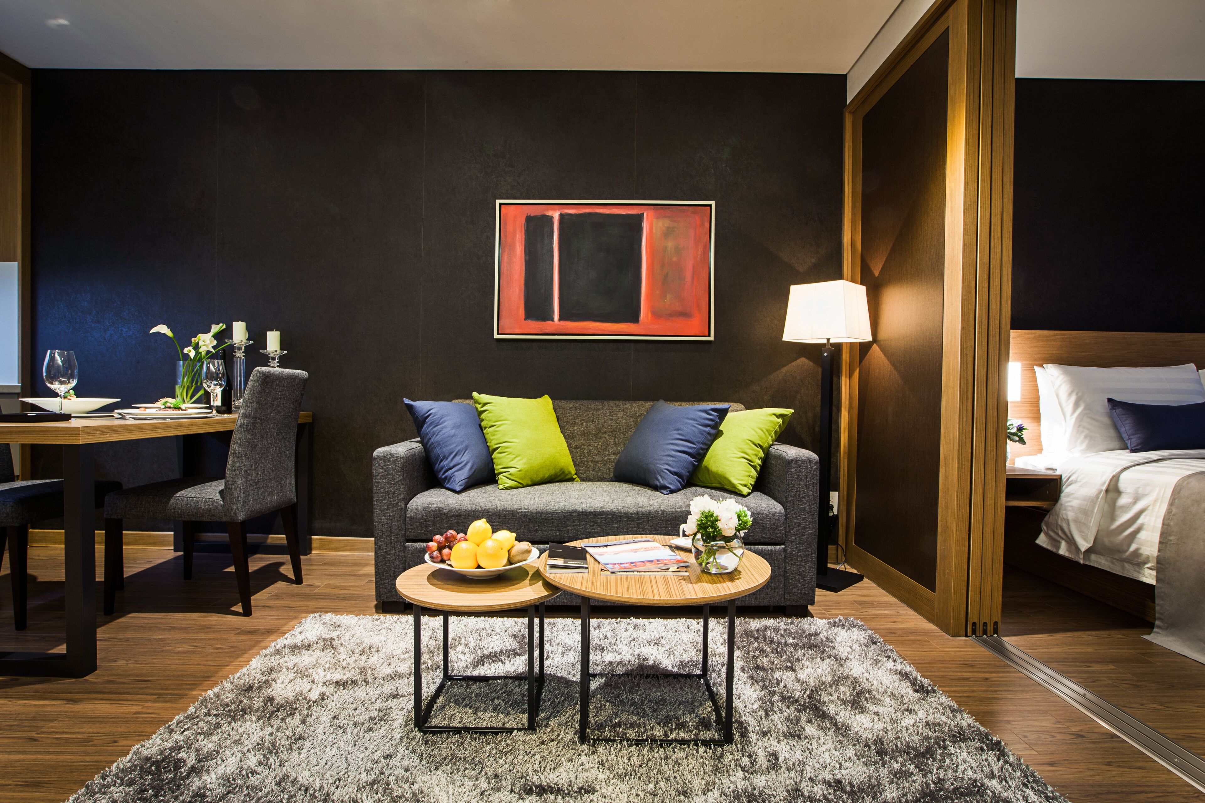 Habitación de hotel moderna con pared oscura, un cuadro abstracto enmarcado, sofá gris con cojines verdes y azules, una mesita de centro redonda con frutas, una mesita auxiliar con un florero, un escritorio de madera con una silla, y una cama con ropa blanca.
