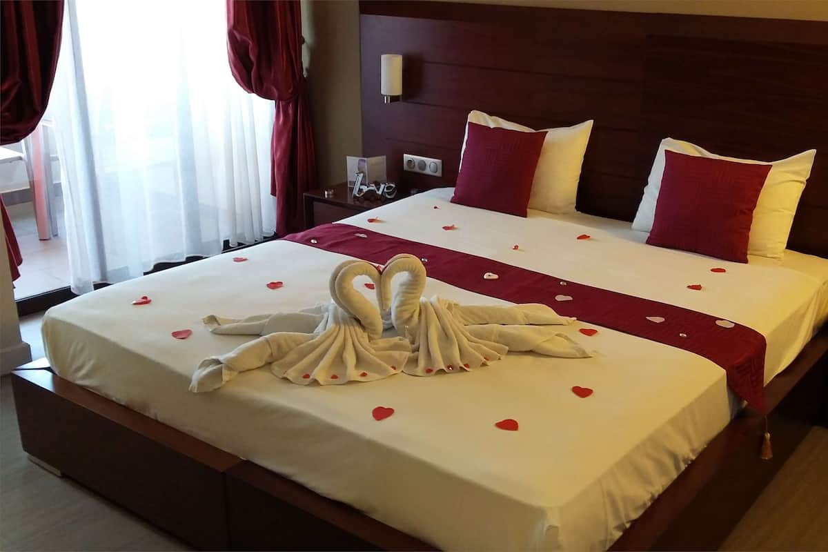 A double bed with a red and white bedspread, two pillows, a red and white throw blanket, and a towel arranged in a swan shape on the bed.