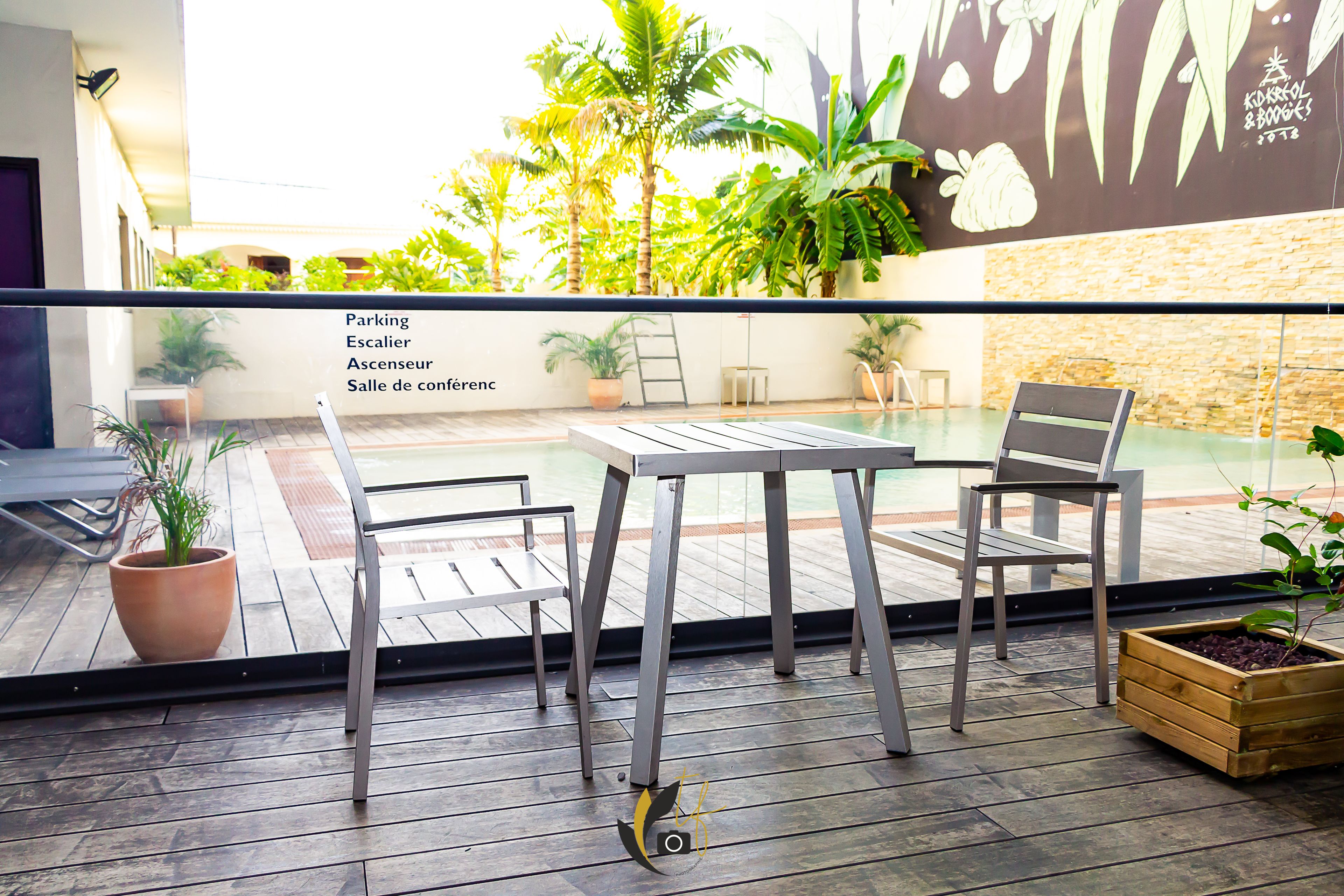 A balcony with a table and chairs, potted plants, and a view of a pool and greenery.