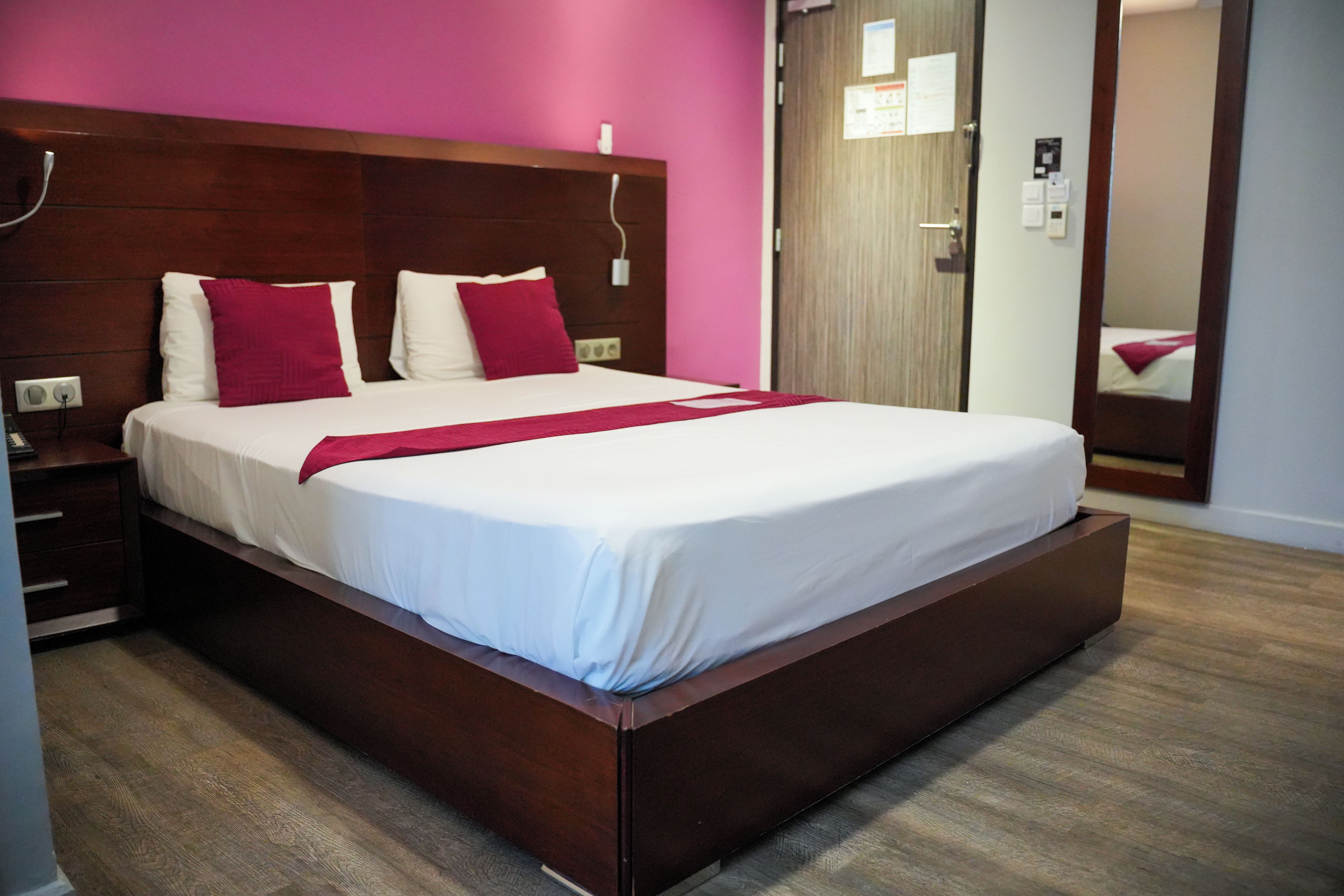 A hotel room with a large bed, wooden headboard, and a pink wall.