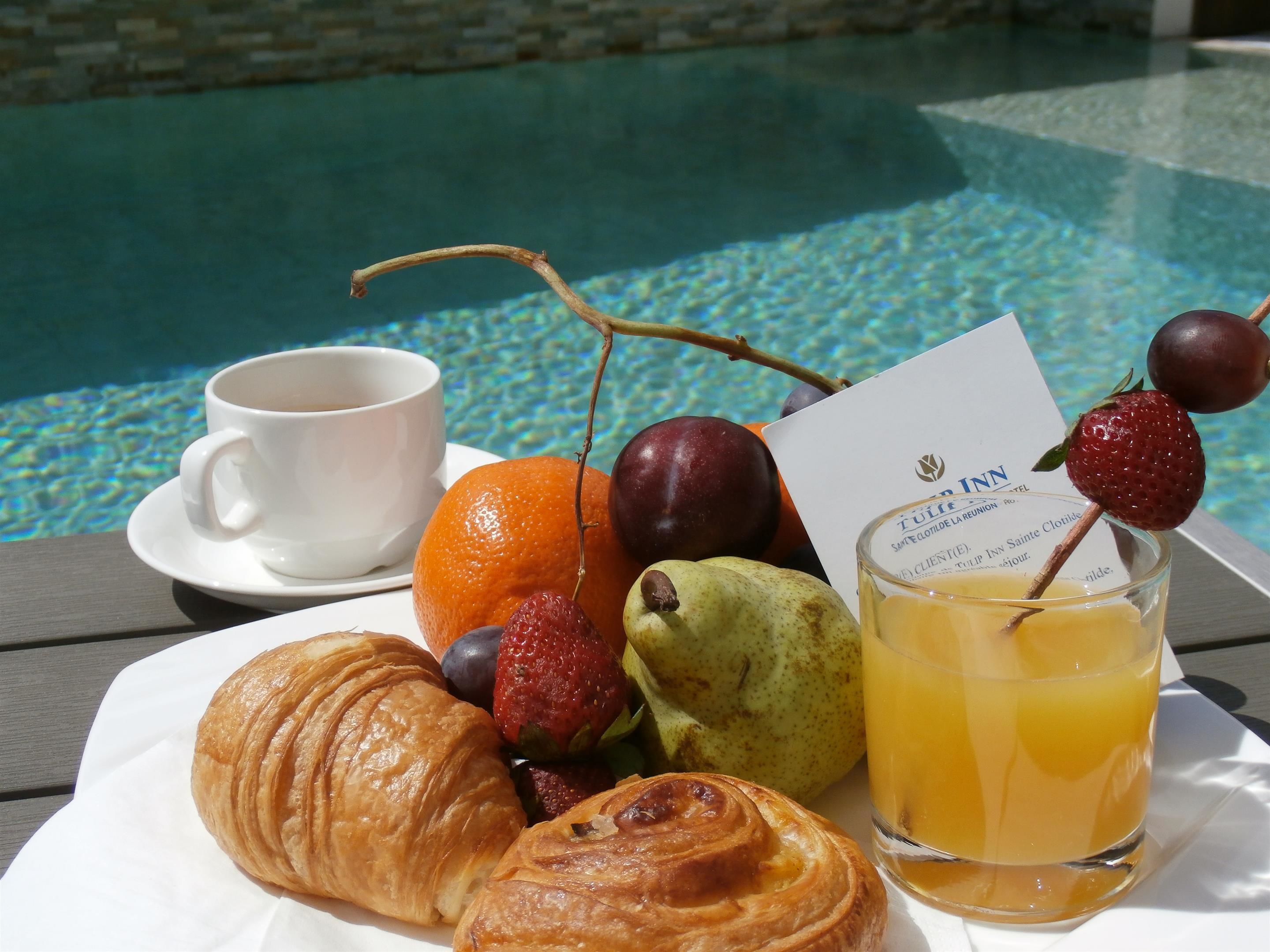 Una bandeja con croissants, frutas, un vaso de zumo de naranja y una taza de café junto a una piscina.