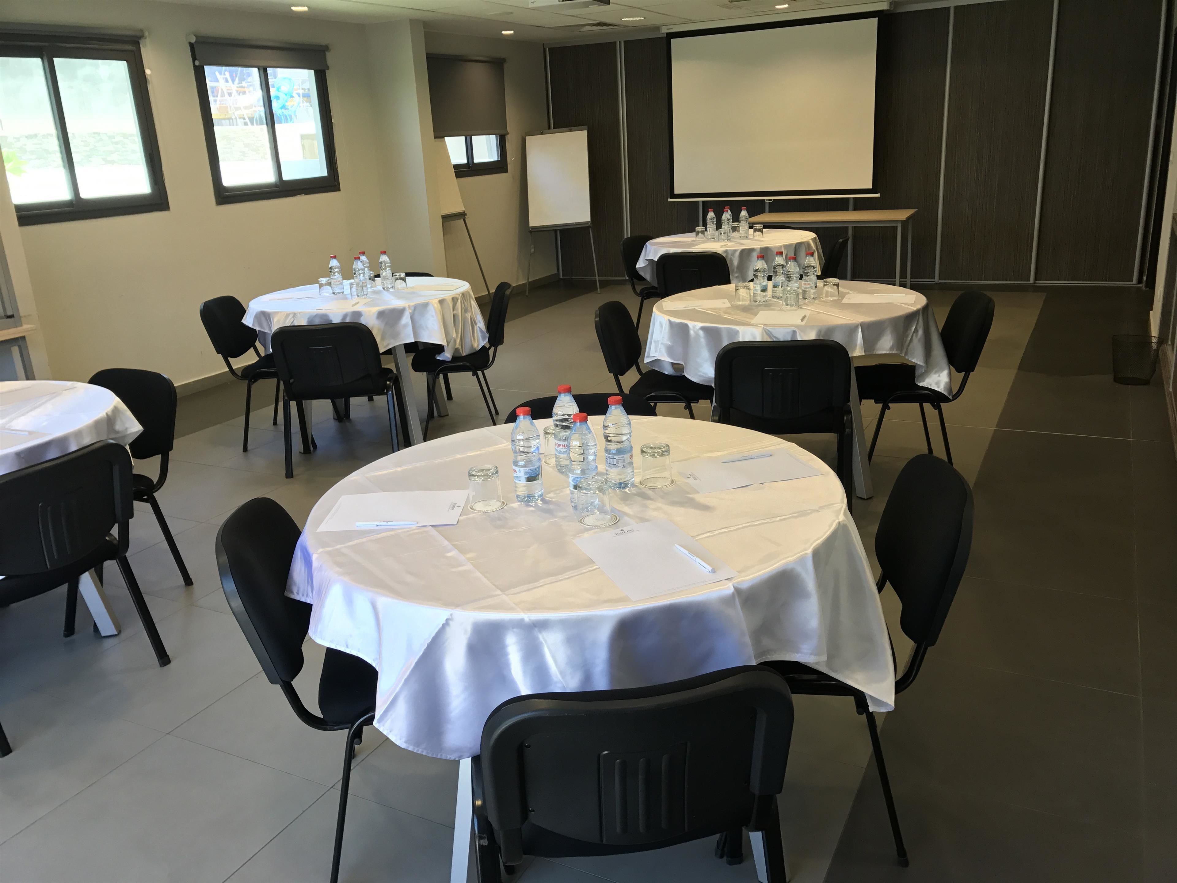 Une salle de conférence dotée de plusieurs tables rondes, chacune recouverte d ’une nappe blanche et équipée de bouteilles d’eau, de verres et de documents.