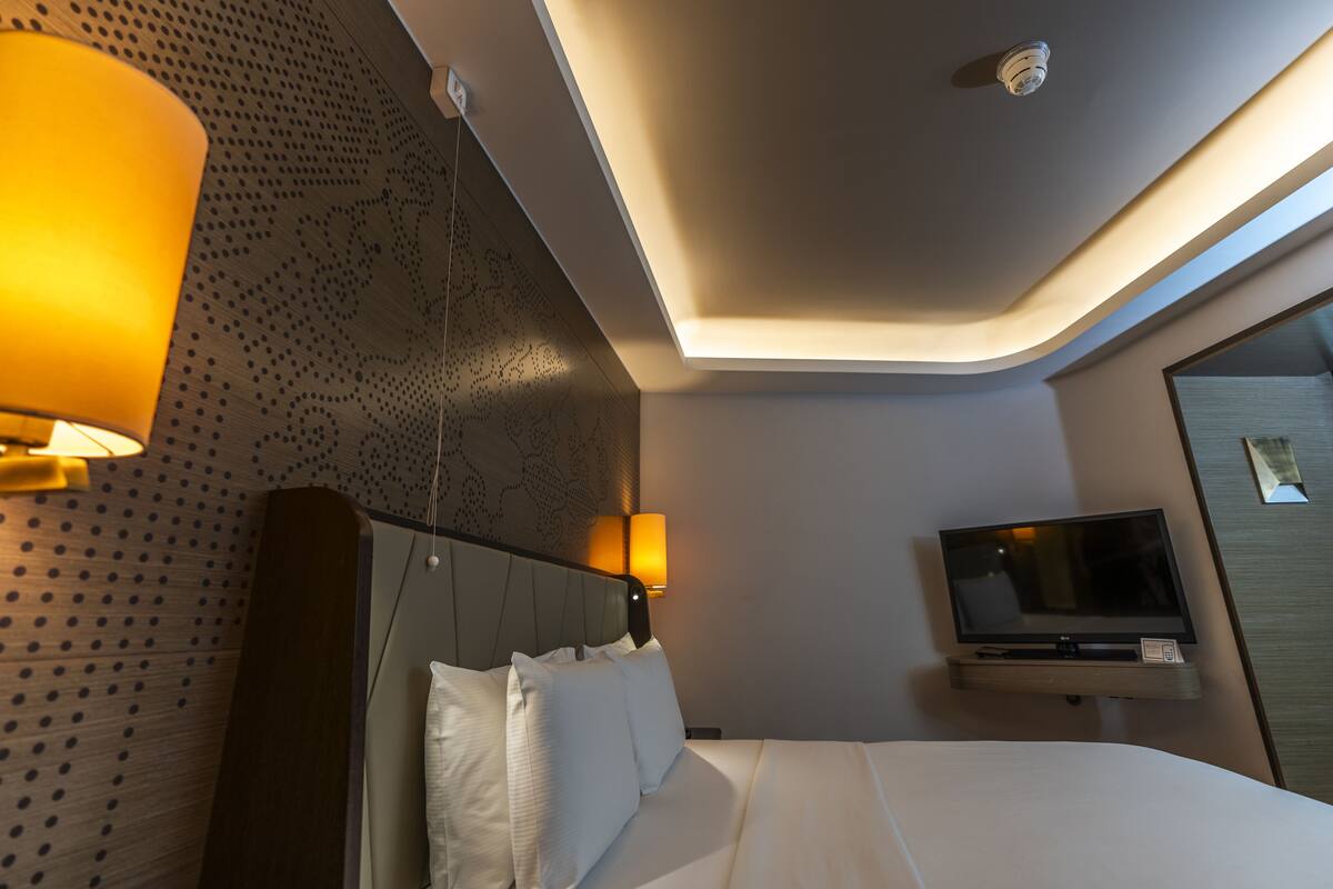 A modern hotel room with a neatly made bed, a flat-screen TV mounted on a wall, and a ceiling light fixture.