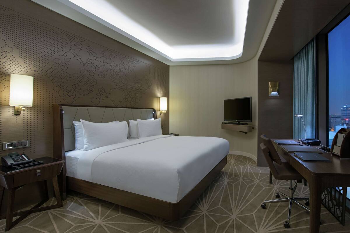 A modern hotel room with a large bed, a desk, a TV, and a city view.