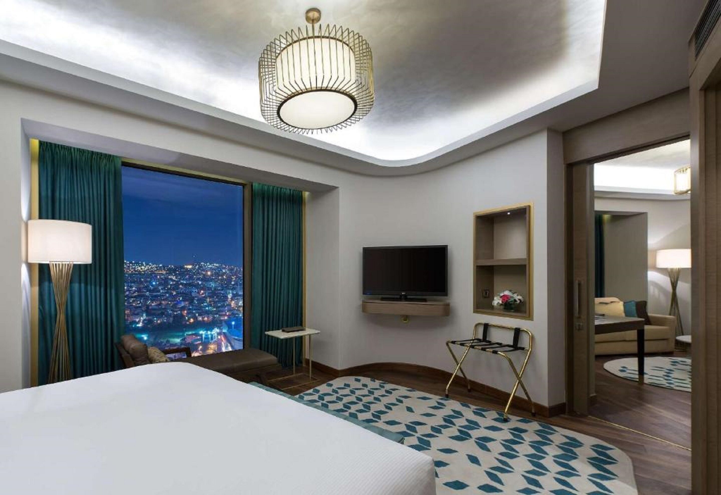 A hotel room with a large bed, a TV, a small table, and a view of the city at night.