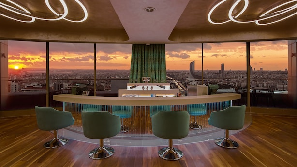 A modern bar with a city view at sunset, featuring a bar counter, four bar stools, and a green curtain. - Hilton Istanbul Kozyatagi (Istanbul)