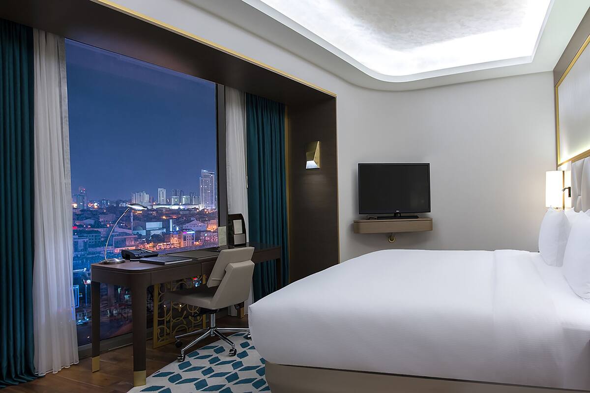 A hotel room with a large bed, a desk with a chair, a television, and a view of a cityscape at night.