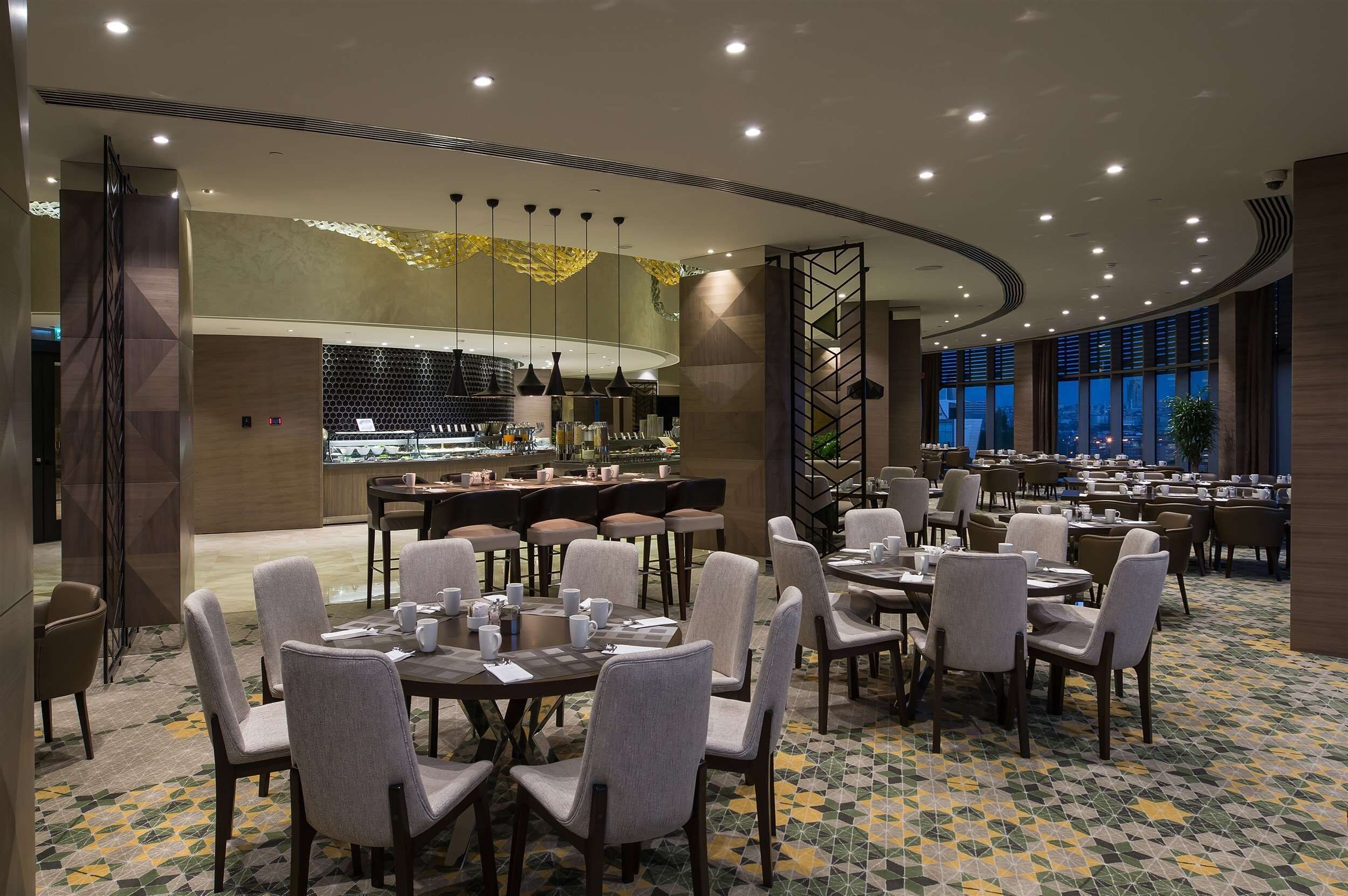 A modern dining area with round tables and chairs, a bar area, and large windows offering a city view.