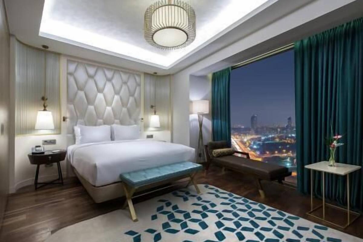 A hotel room with a large bed, a bench, a view of the city at night, and a skylight.