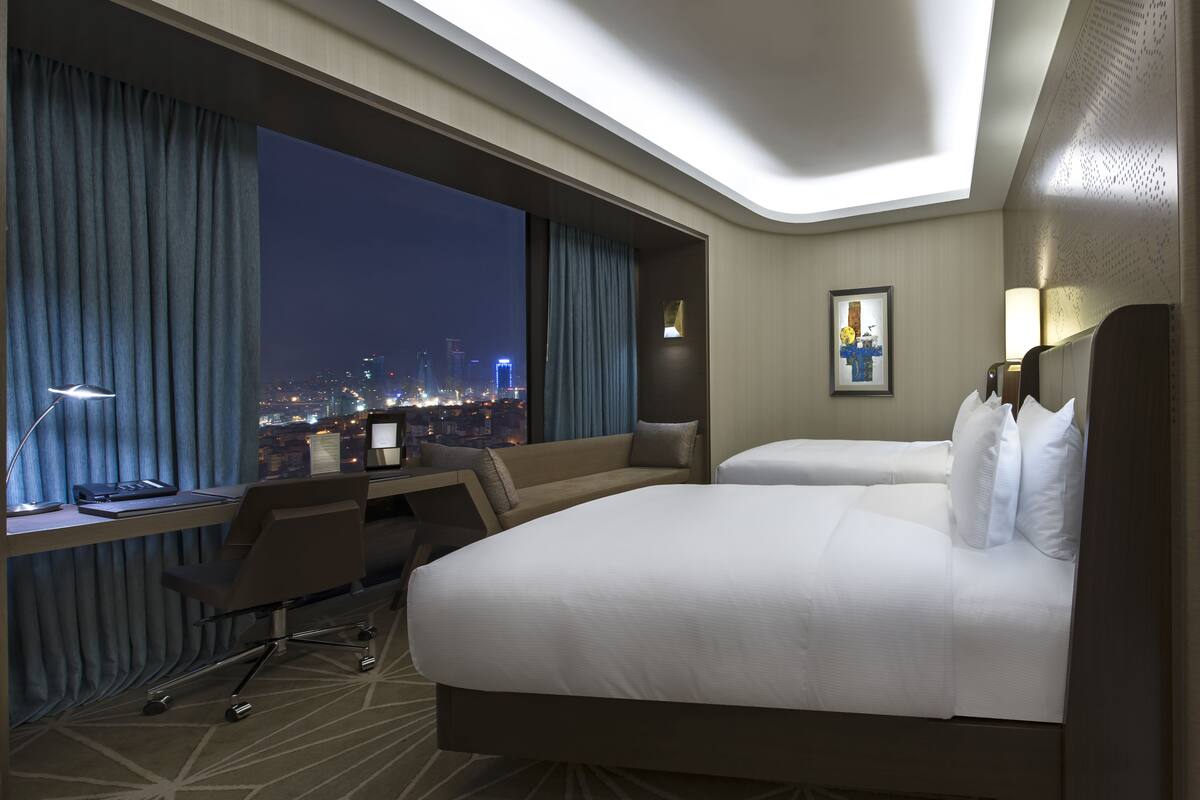 A hotel room with two beds, a desk, and a chair. There is a large window with curtains, a city view, and a framed picture on the wall.