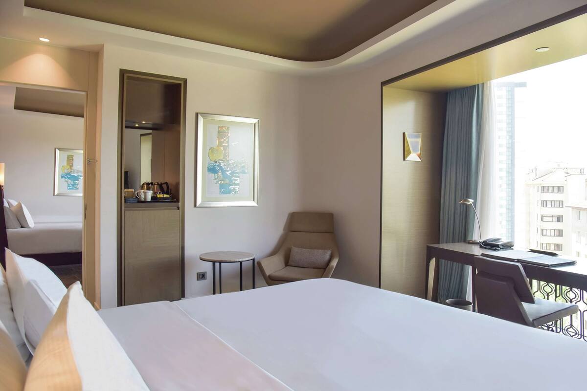 A hotel room with a large bed, a desk with a chair, a small table, and a chair. There are framed pictures on the wall and a window with a view of buildings.
