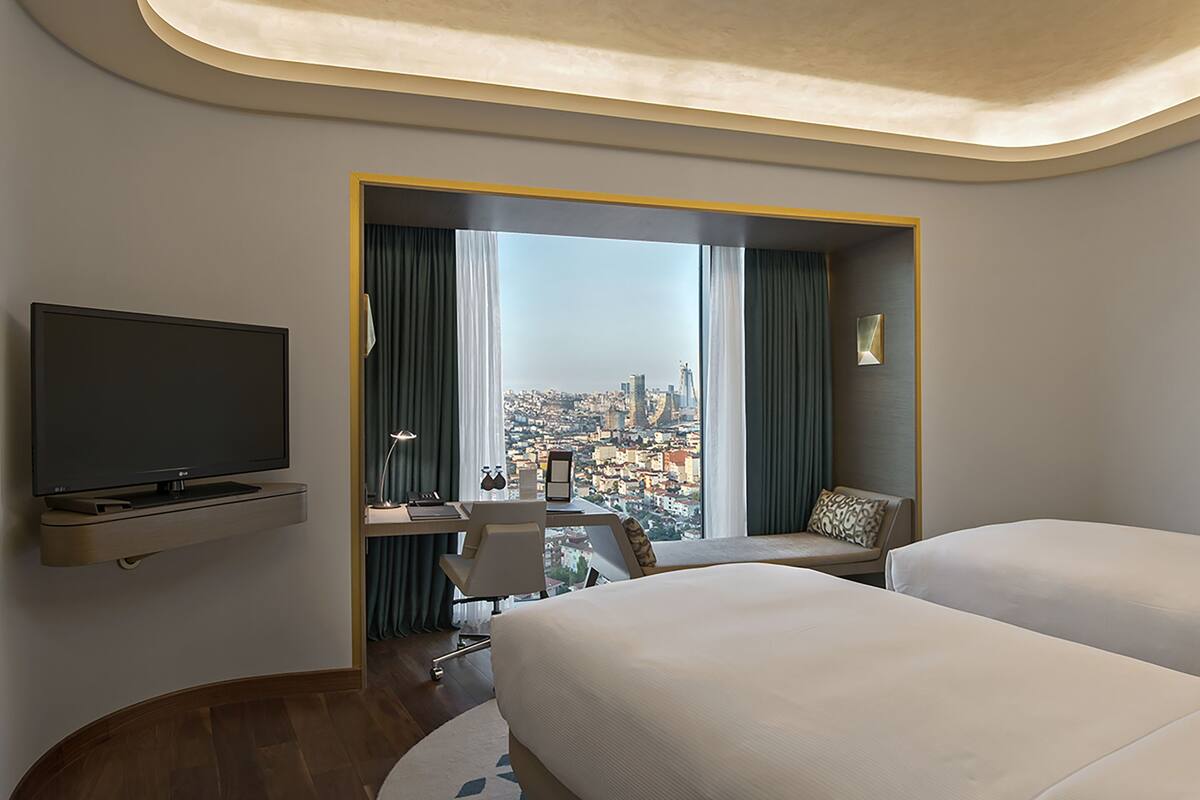 A hotel room with a bed, a desk with a chair, a television, and a large window offering a city view.