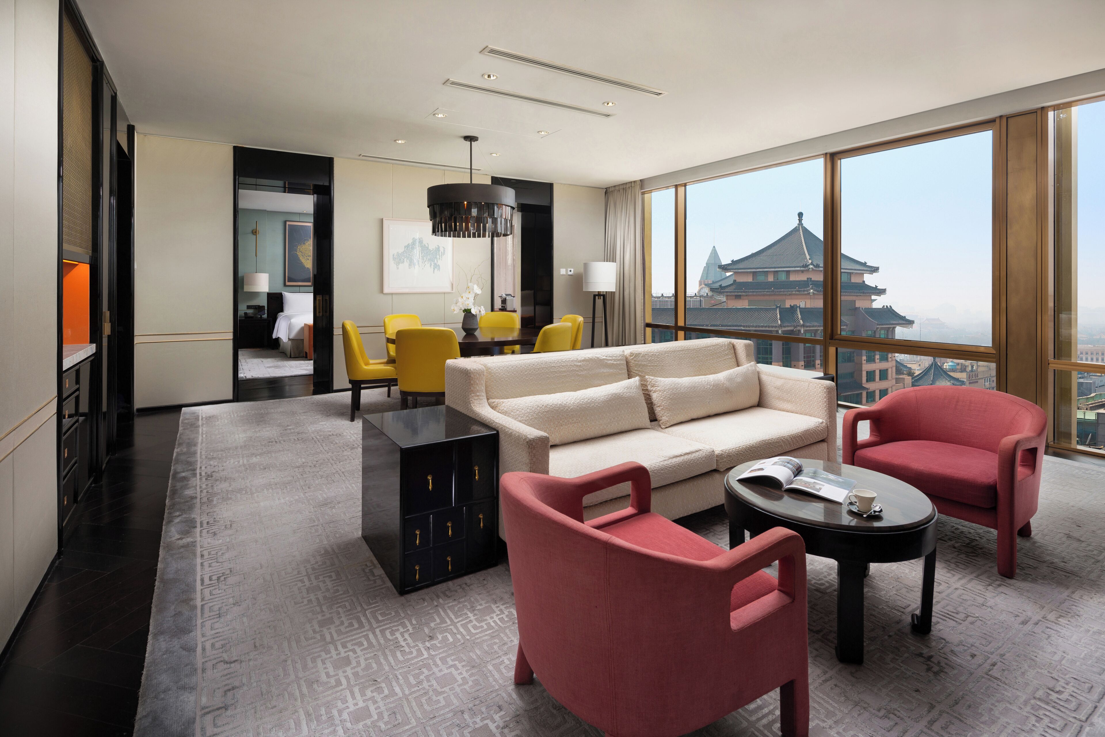 Een moderne hotelkamer met een groot raam, een beige bank, gele stoelen, een rode fauteuil, een zwart bijzettafeltje en een decoratieve lamp.