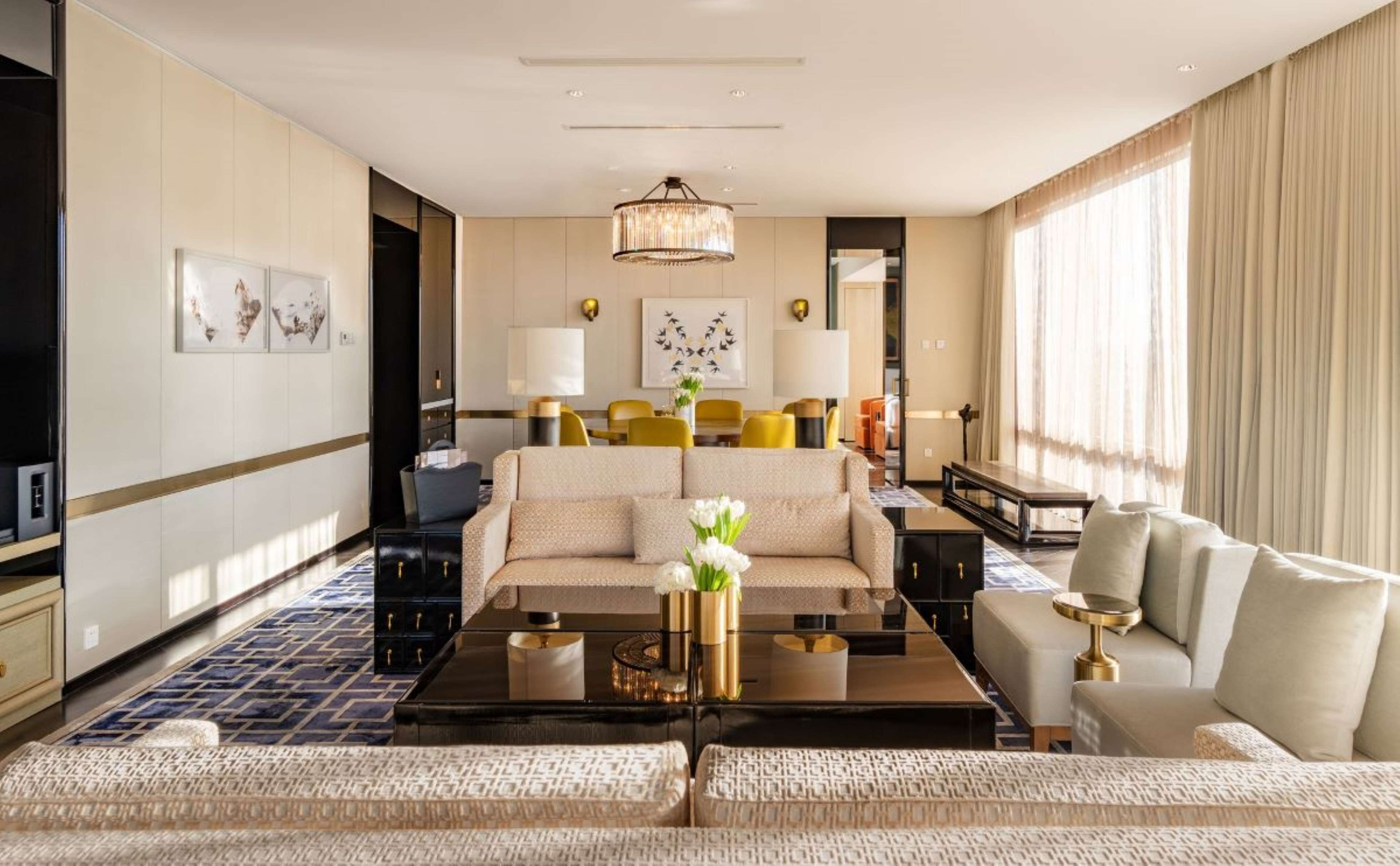 A modern hotel room with a sofa set, a dining area, and a large window with curtains.