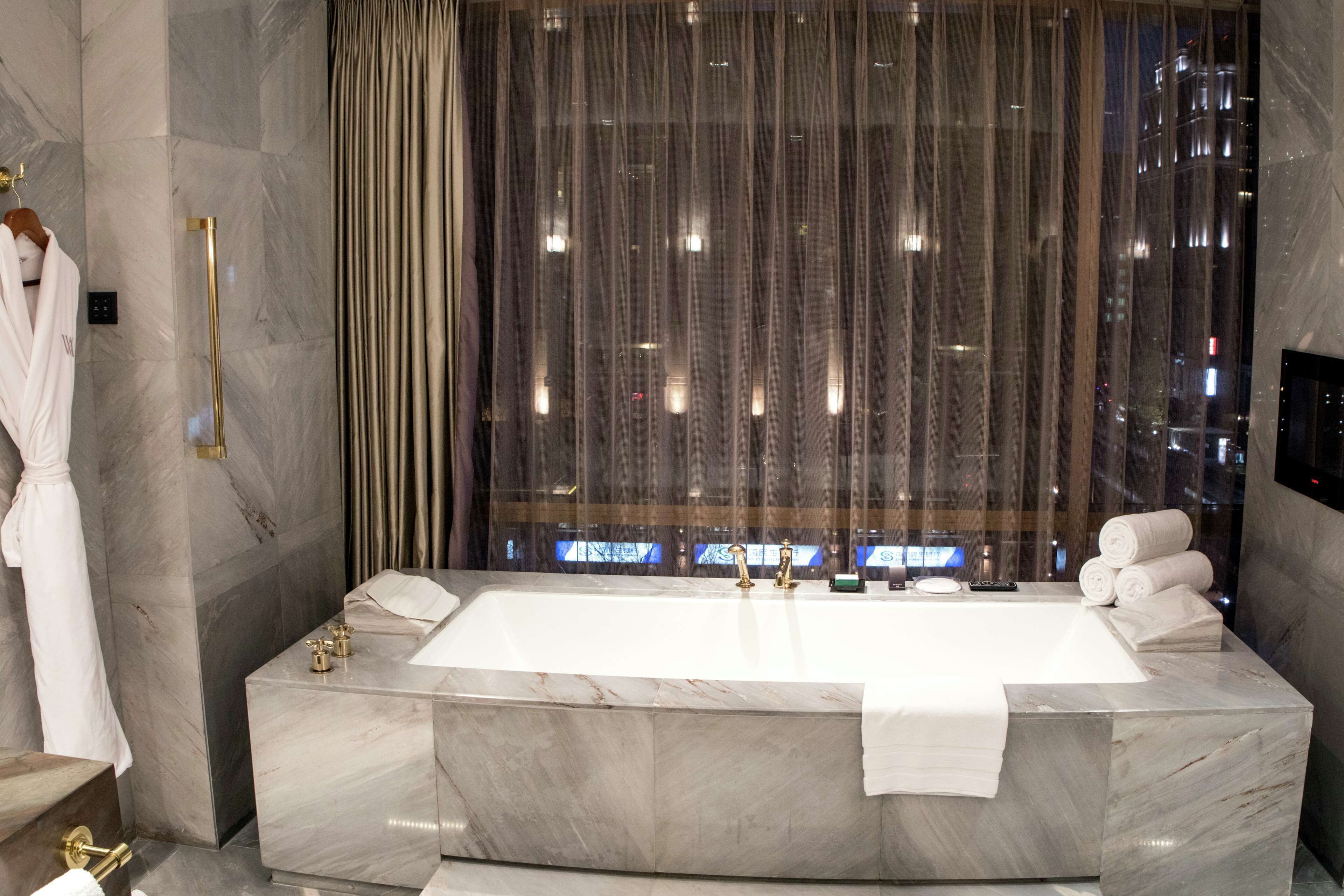 A modern bathroom with a large marble bathtub, a glass-enclosed shower, and a view of the city at night.