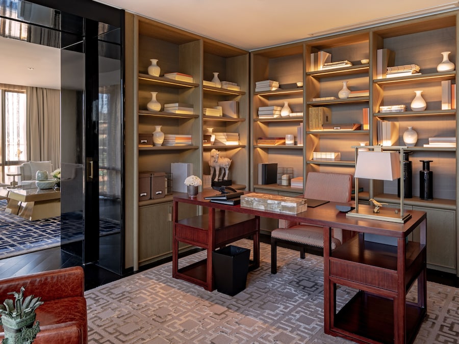 Habitación de hotel moderna con una amplia estantería, un escritorio con lámpara, un sillón de cuero y una alfombra estampada.
