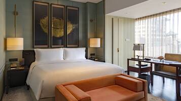A hotel room with a large bed, two bedside tables with lamps, a sofa, a desk with a chair, and a large window with curtains.
