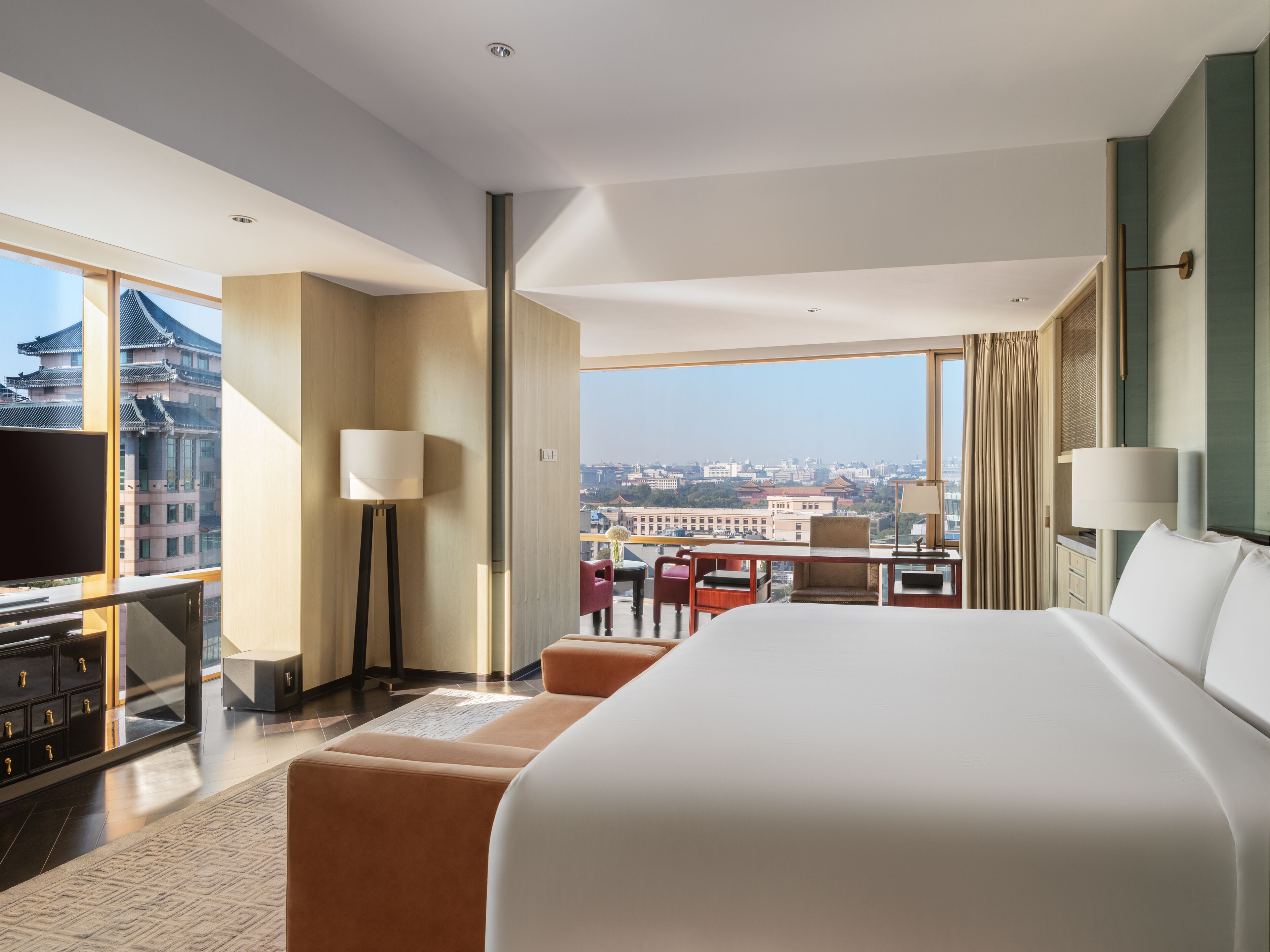 A modern hotel room with a large bed, a desk with a chair, a TV, and a view of a cityscape.