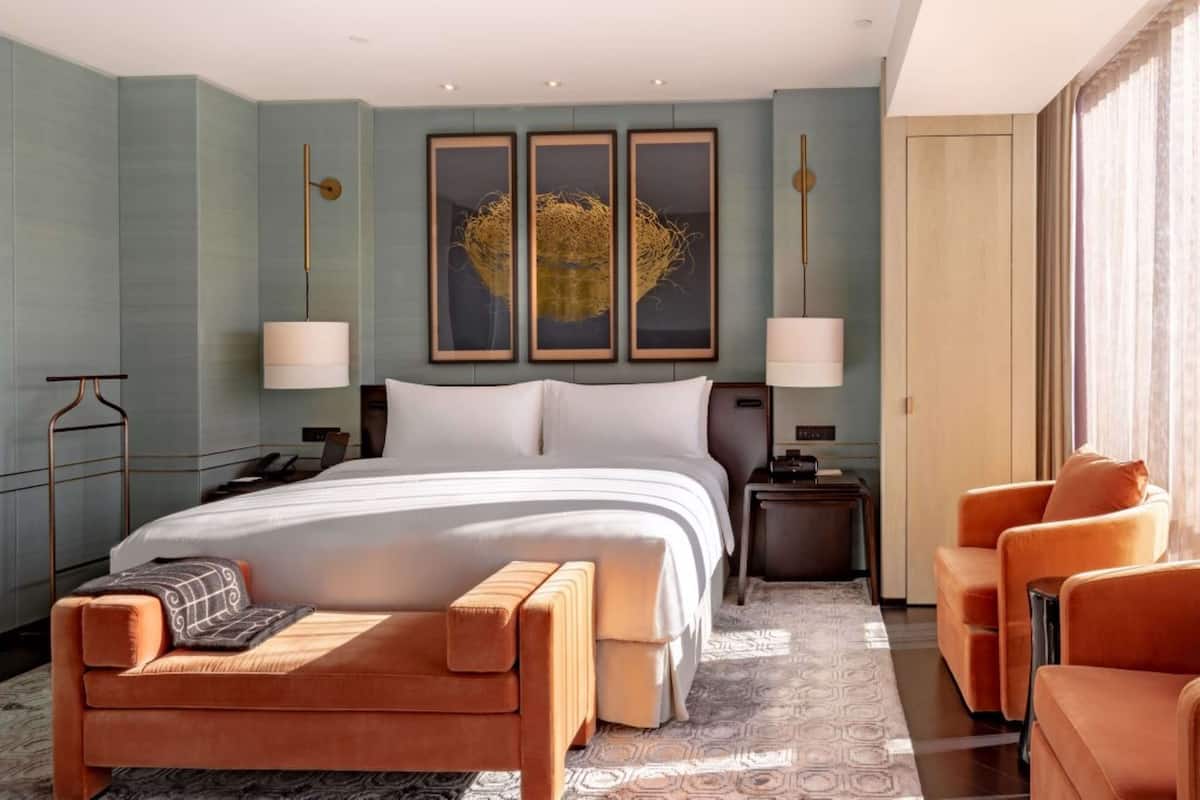 A modern hotel room with a large bed, a seating area with a sofa and armchairs, a desk, and a large window with curtains.