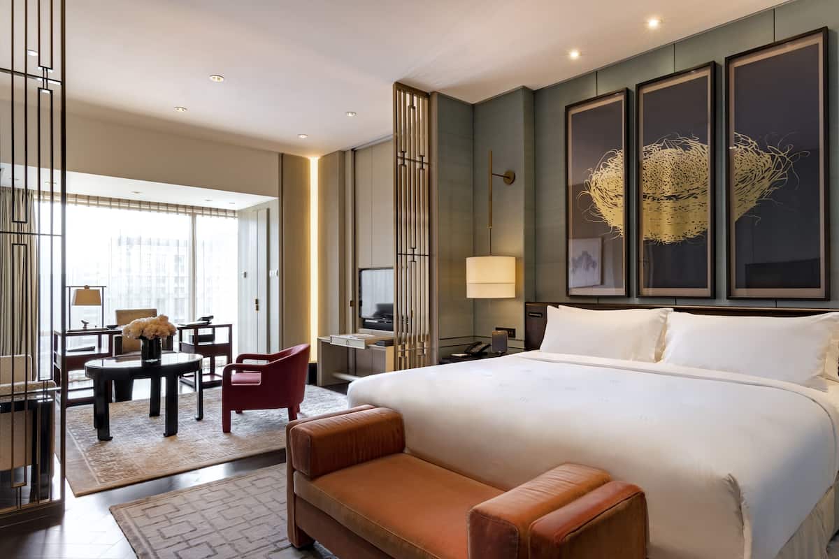 A modern hotel room with a large bed, a seating area with a table and chairs, a desk with a computer, and artwork on the walls.