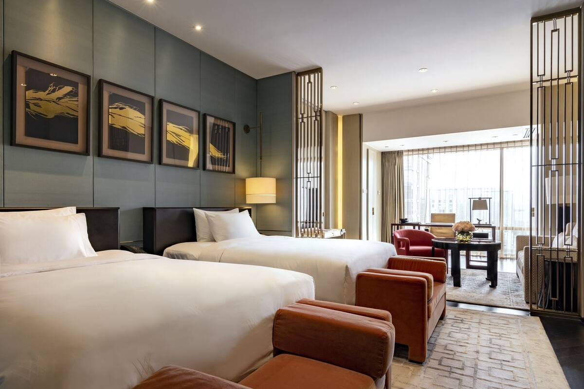 A hotel room with two beds, a desk, a chair, and a view of the city.