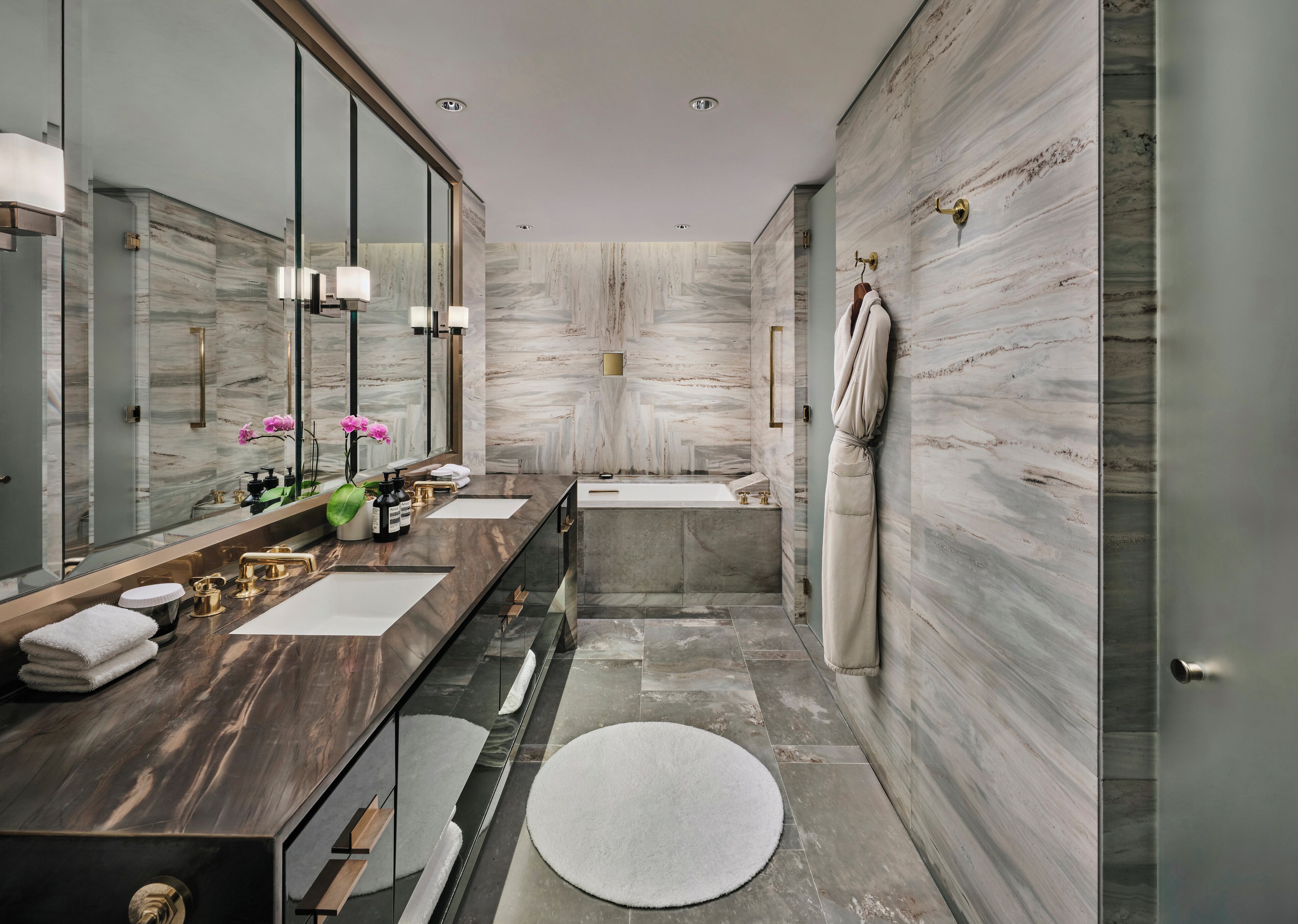 A modern bathroom with a marble wall, a large mirror, a dual sink vanity, a bathtub, and a round rug on the floor.