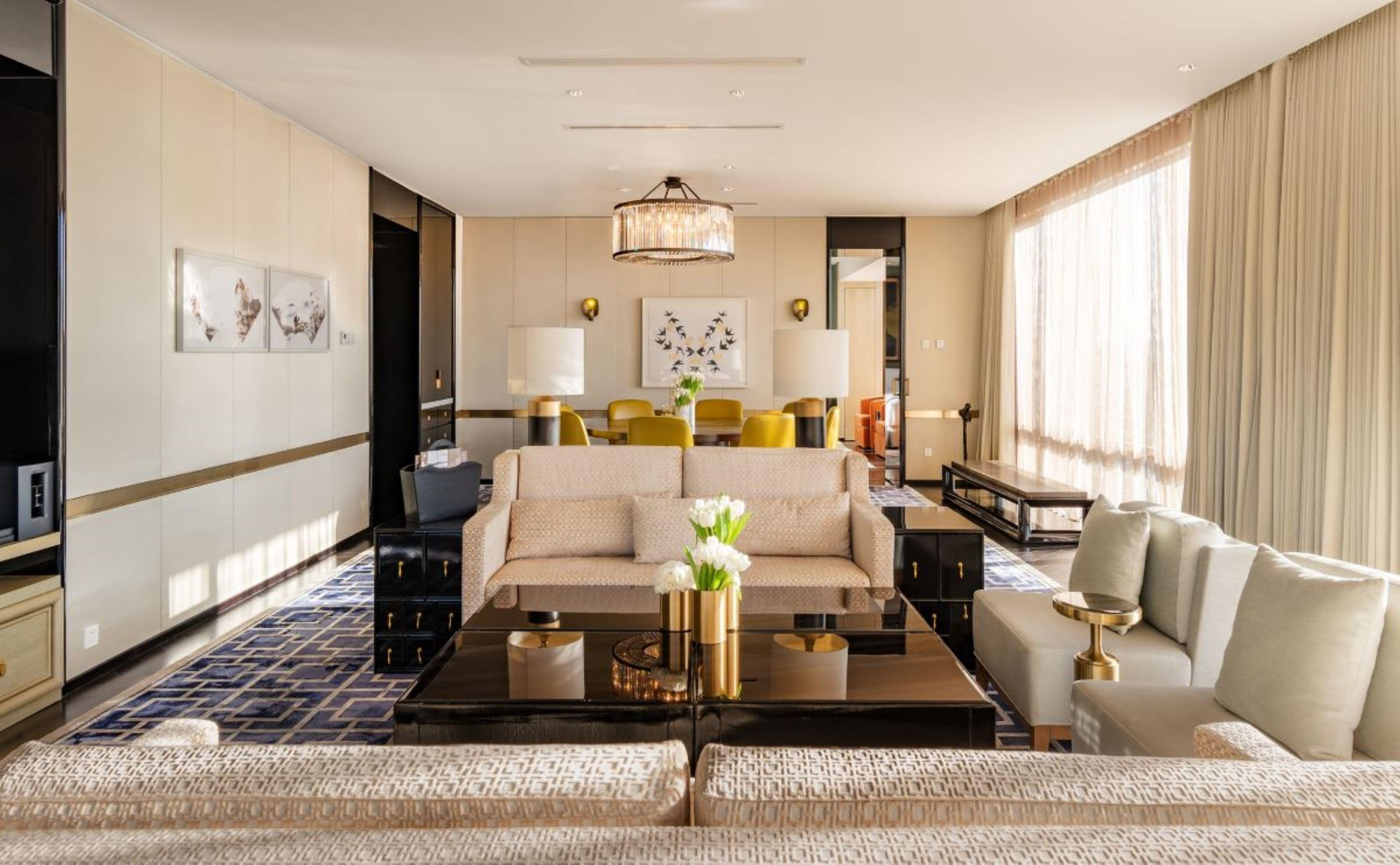 A hotel room with a sofa, armchairs, a coffee table, a chandelier, artwork, and a dining area.