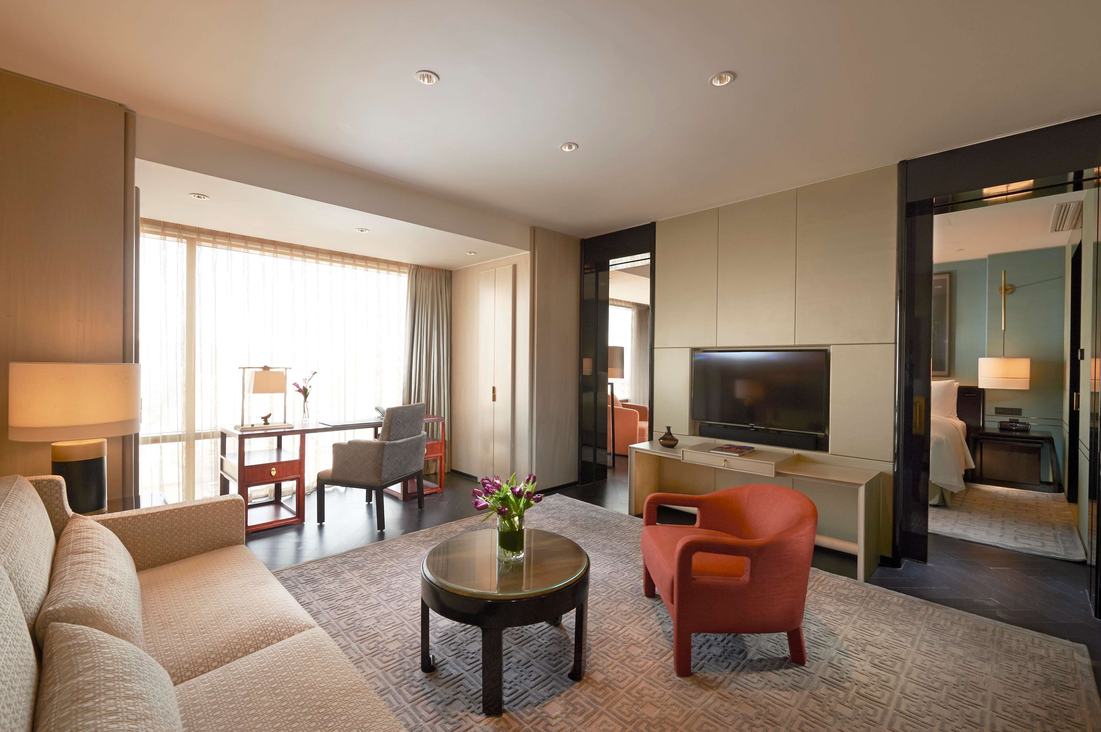 Une chambre d’hôtel moderne, dotée d’un canapé, d’un fauteuil, d’une table basse, d’une télévision et offrant une vue sur la ville grâce à de grandes fenêtres.