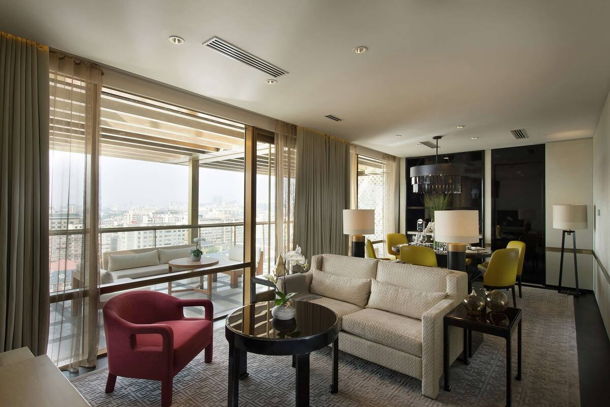 A modern hotel room with a large window, a sofa, a red chair, a small table, and a dining area with yellow chairs and lamps.