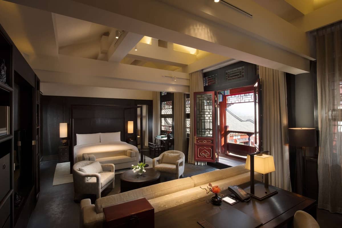 A hotel room with a bed, sofa, armchair, and a small table with a lamp. There is a view of a traditional building outside the window.