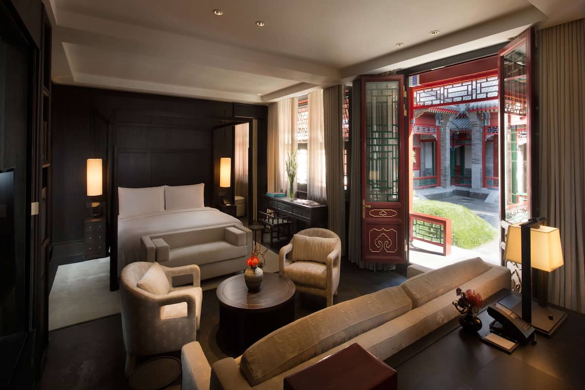 A modern hotel room with a large bed, a sofa, a coffee table, and a desk. There is a view of a traditional courtyard through an open door.