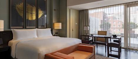 A modern hotel room with a large bed, a sofa, a desk with a chair, and large windows offering a city view.