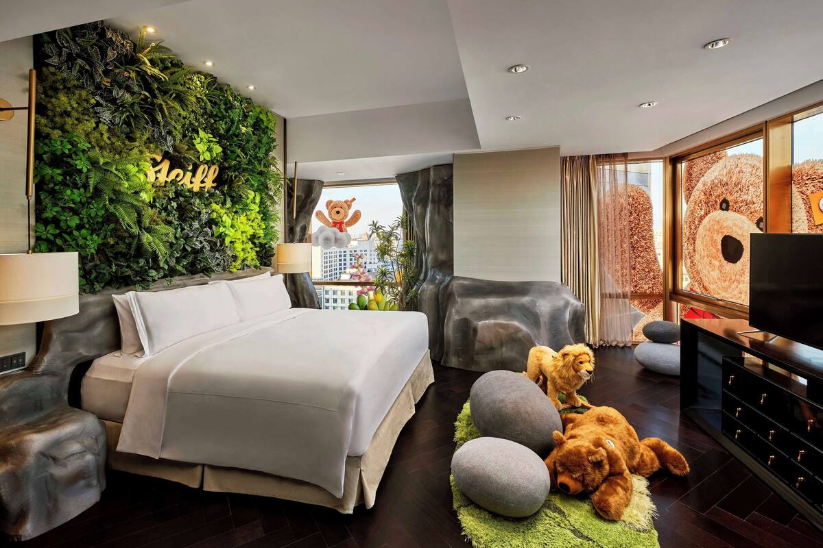 A modern hotel room with a large bed, a green wall with plants, a teddy bear, and a TV.