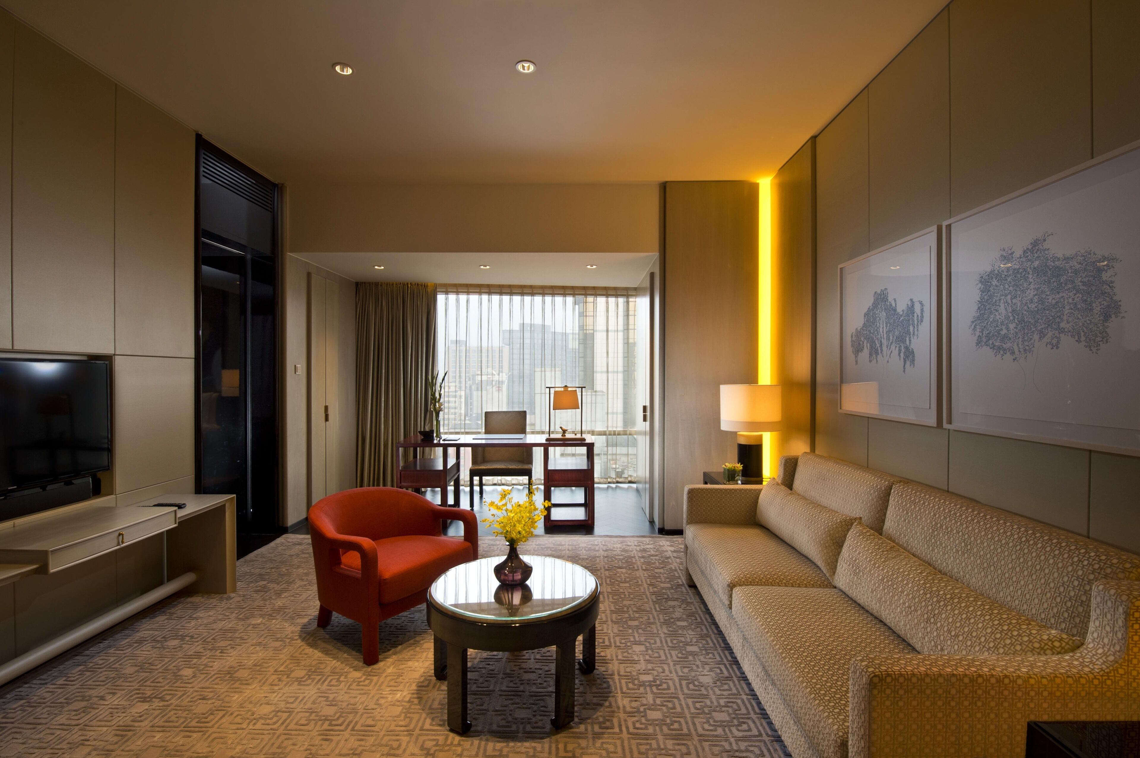 A hotel room with a TV, a sofa, a red armchair, a coffee table with flowers, and a dining area with a table and chairs.