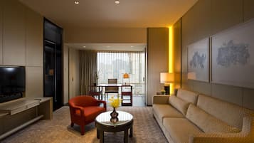 A hotel room with a TV, a sofa, a red armchair, a coffee table with flowers, and a dining area with a table and chairs.