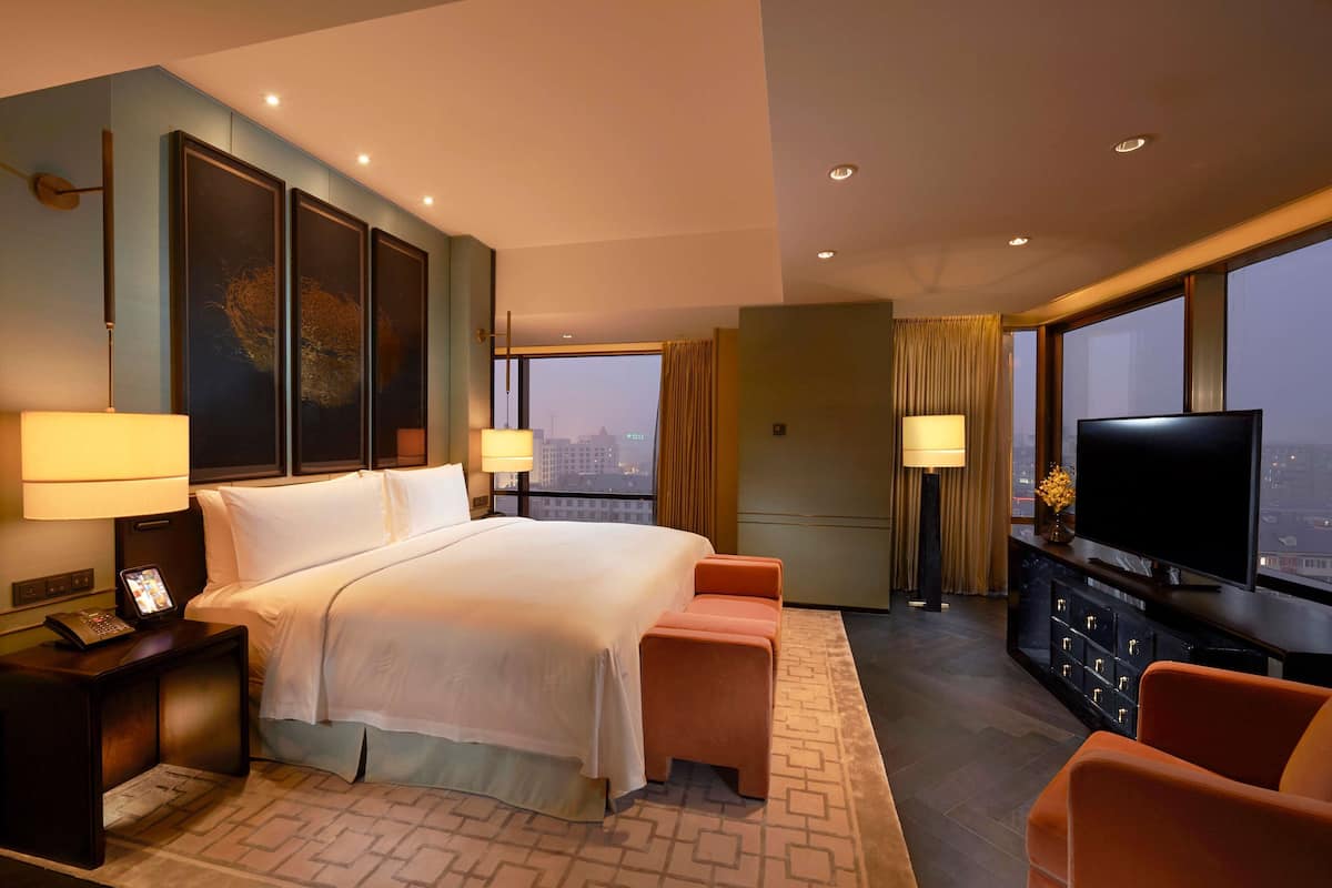 A hotel room with a bed, bedside lamps, a TV, a seating area, and a view of the cityscape.