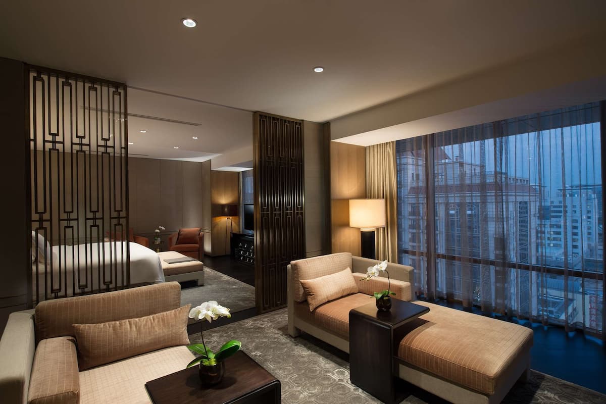 A modern hotel room with a bed, two sofas, a coffee table, and a large window with a city view.