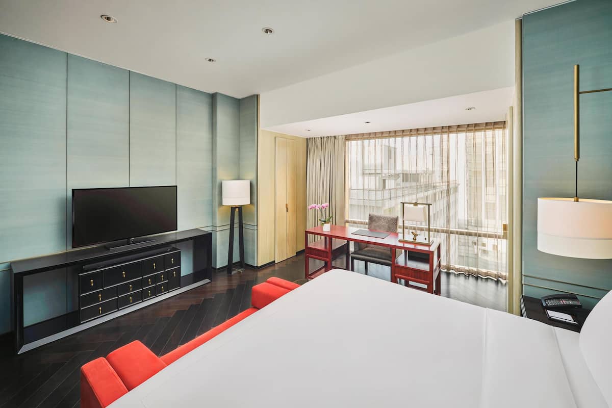 A hotel room with a large bed, a TV on a black console, a desk with a lamp, and a view of the city through large windows.