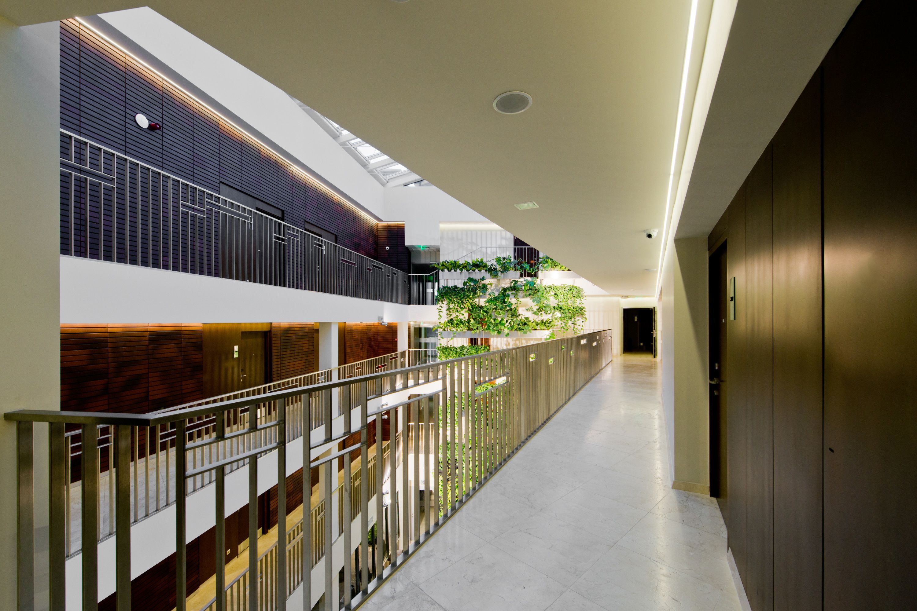A modern hotel corridor with a glass railing, a staircase, and a view of a green wall with plants.