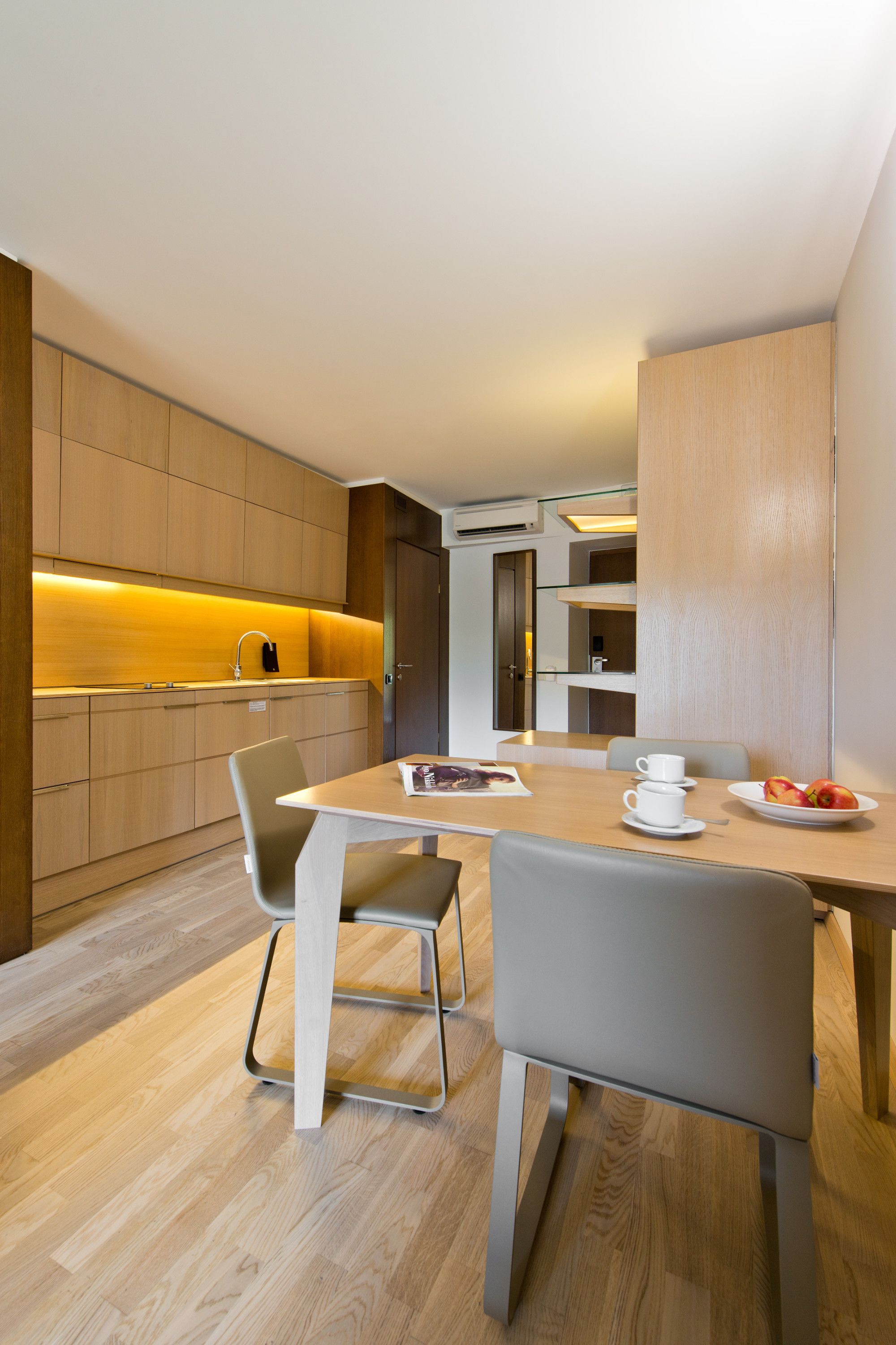 A modern kitchen with wooden cabinets, a dining table with chairs, and a breakfast setting with fruit and a cup.