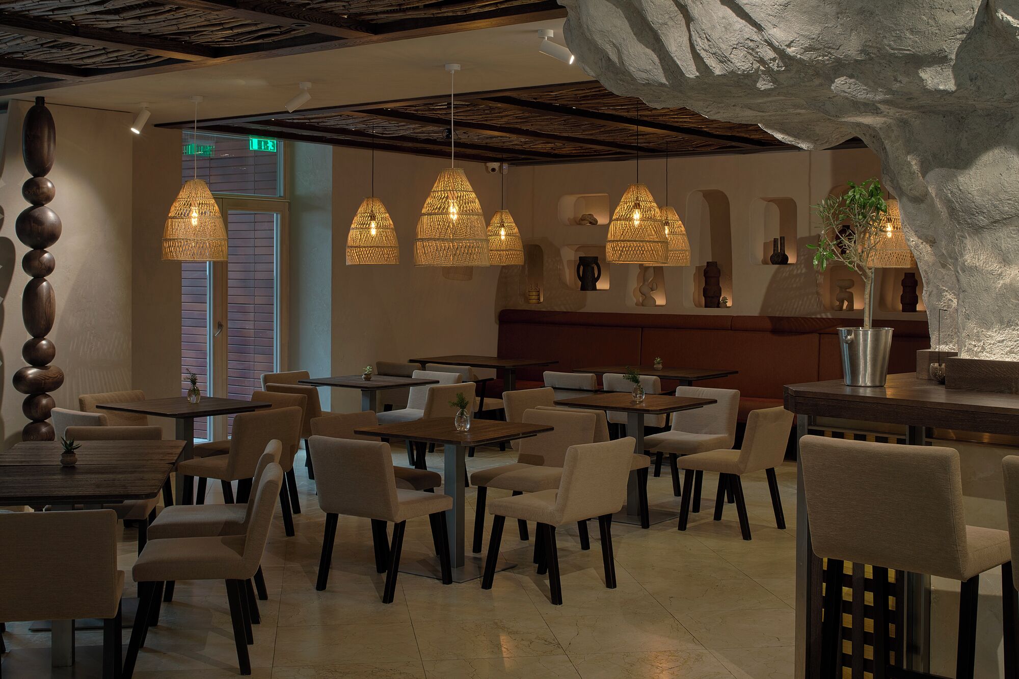 Interior de restaurante con mesas redondas, sillas acolchadas y lámparas de ratán colgantes.