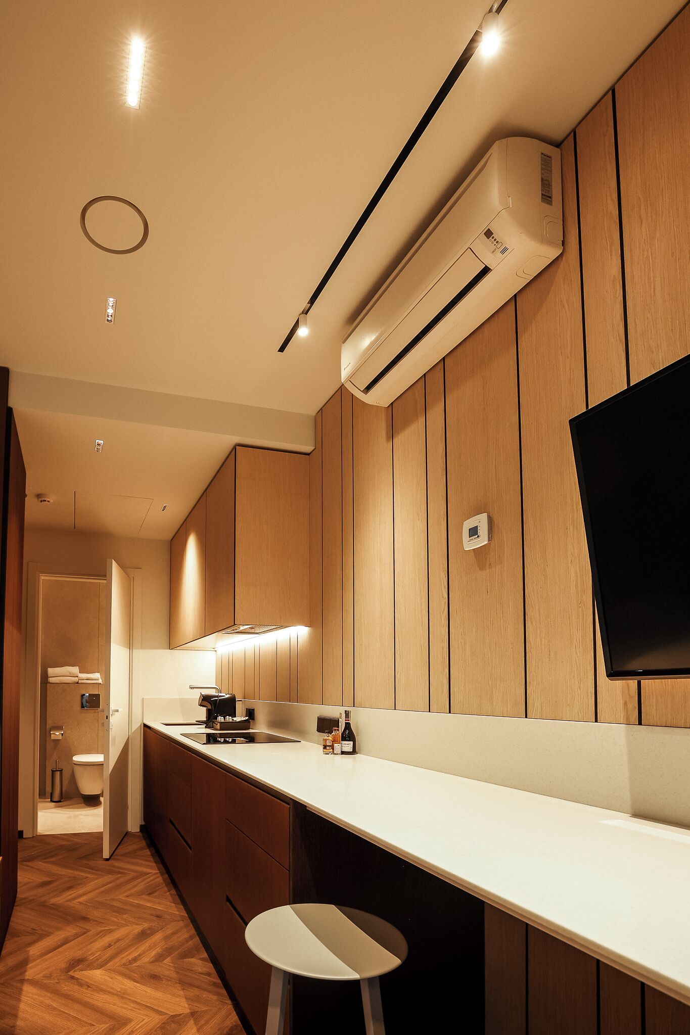 A modern kitchen with wooden paneling, a built-in air conditioner, a flat-screen TV, and a white countertop.