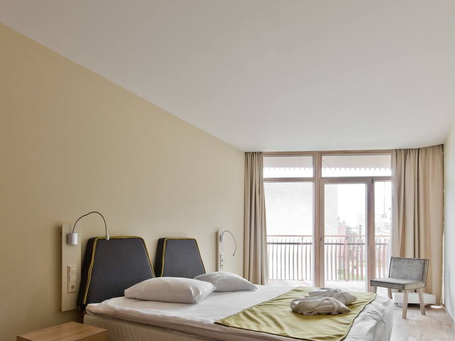 Habitación de hotel moderna con una cama grande, una mesita de noche, una lámpara y vistas al balcón exterior.