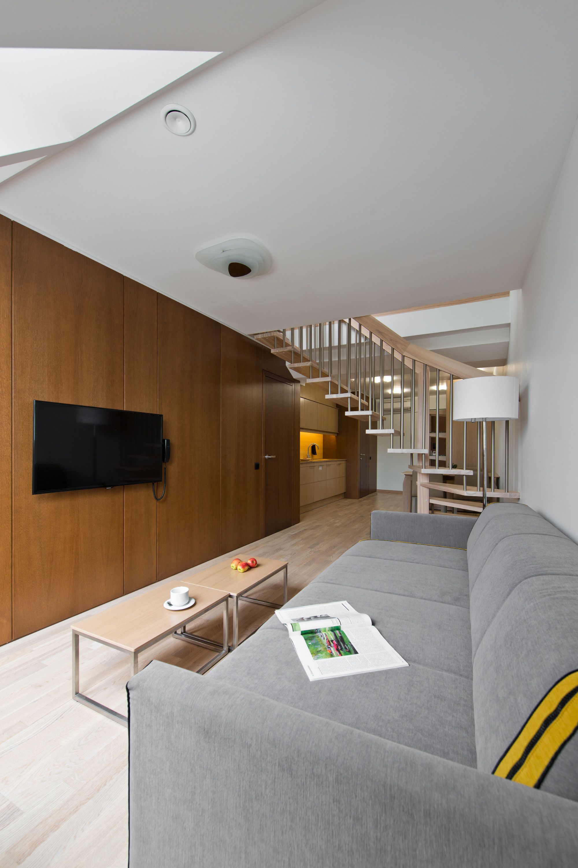 Una sala de estar moderna con un televisor montado en una pared de madera, un sofá gris, una mesa de centro de madera y una escalera que conduce a un nivel superior.