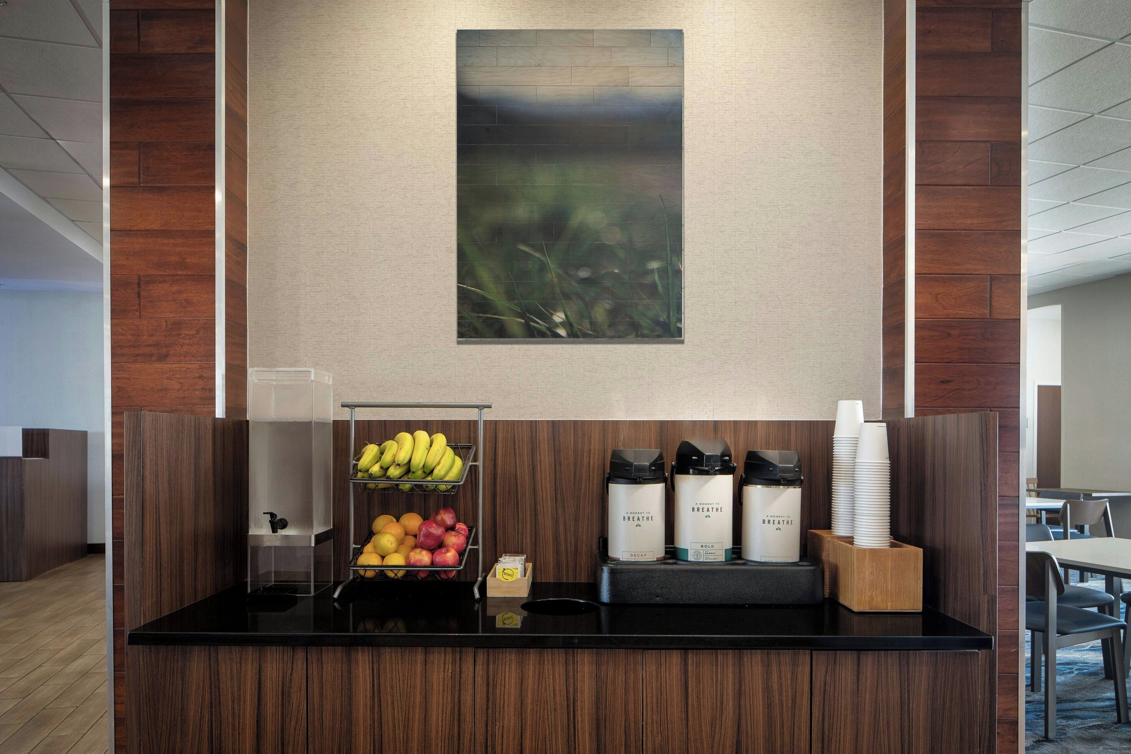 A reception area with a fruit display, coffee dispensers, and a wall-mounted painting.