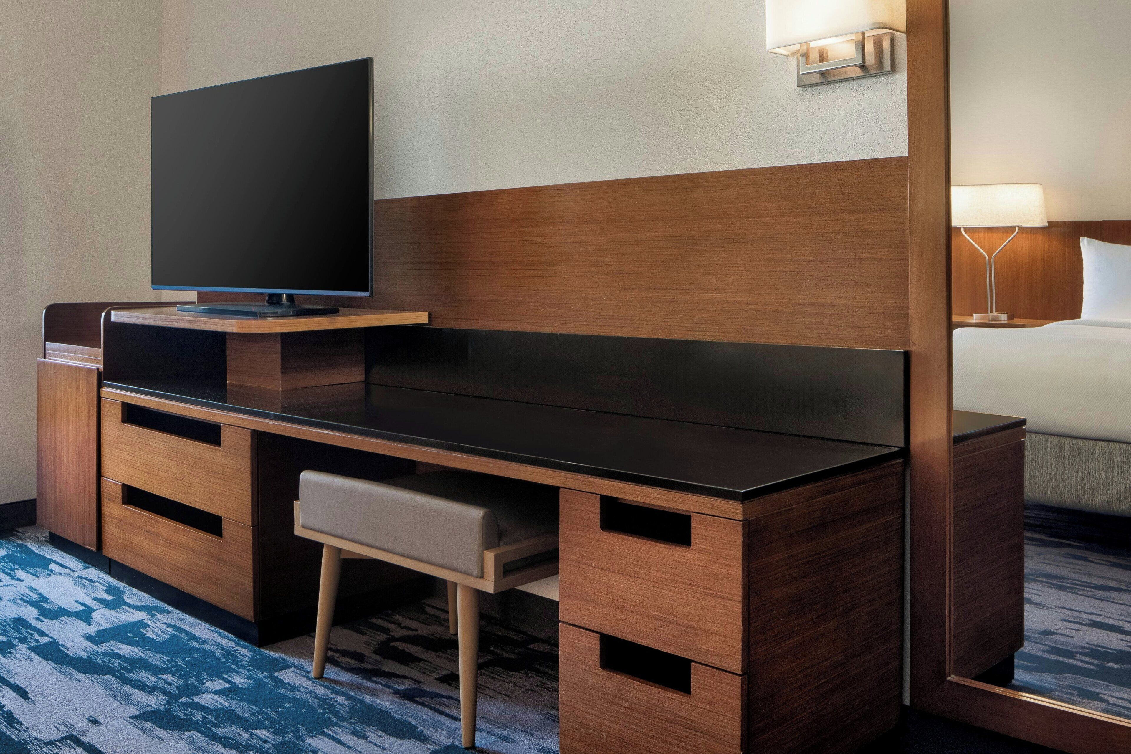 A hotel room with a wooden desk, a television, a bed, and a nightstand with a lamp.