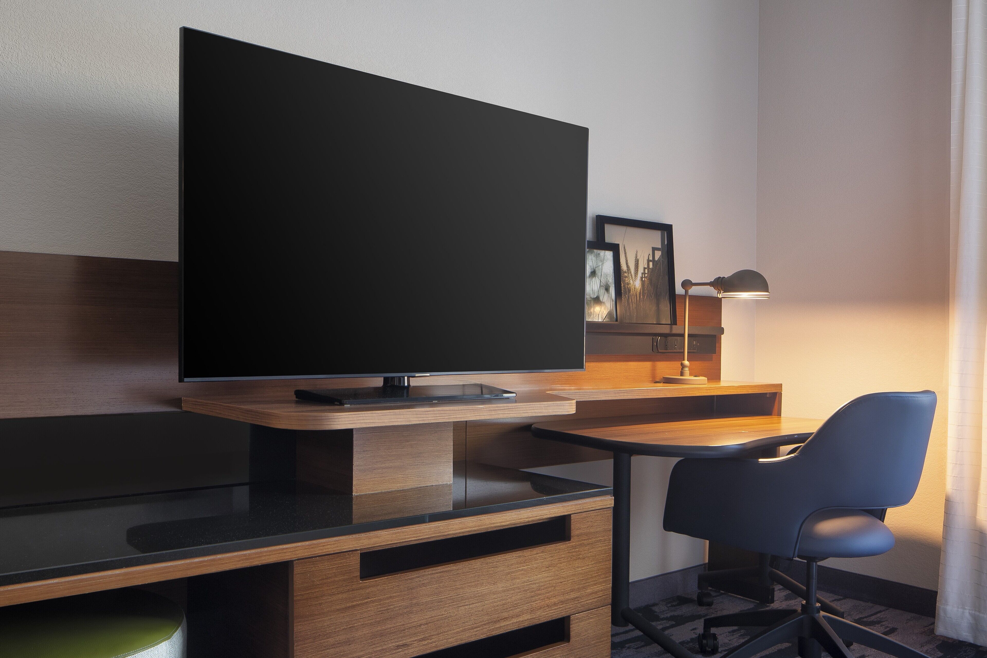 A modern hotel room with a flat-screen TV mounted on a wooden wall, a desk with a chair, and a bedside table with a lamp.