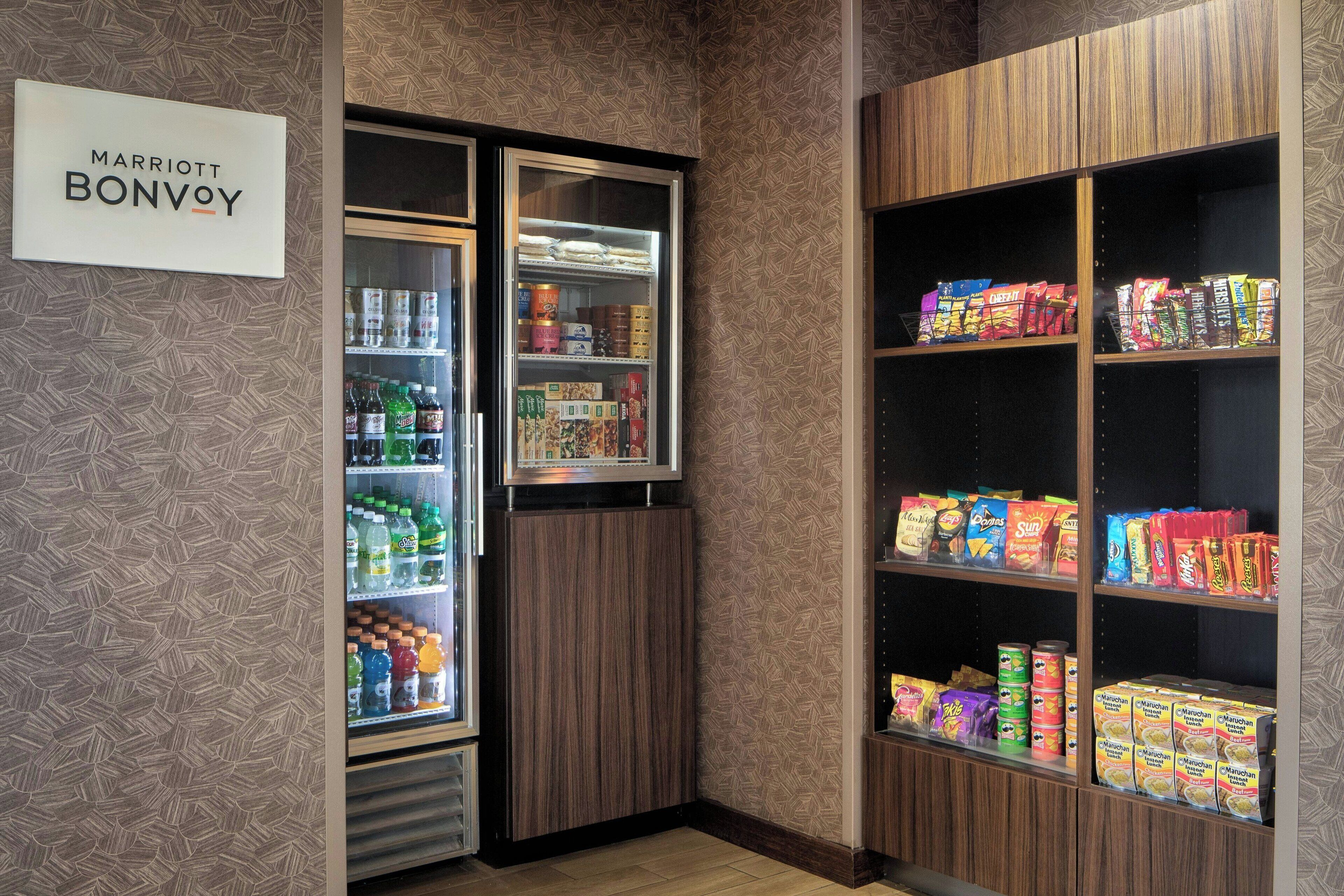 Un minibar con refrigerador lleno de bebidas y aperitivos, y estantes con alimentos envasados.